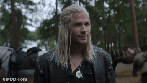 A Man With Long Blonde Hair And A Necklace Is Standing In The Woods . GIF