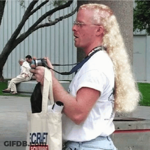A Man With Long Blonde Hair Is Holding A Camera And A Tote Bag That Says Craft On It GIF