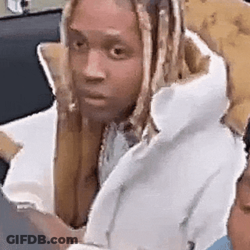 A Man With Long Blonde Hair Is Sitting In A Chair Looking At His Phone . GIF