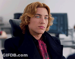 A Man With Long Blonde Hair Is Wearing A Black Coat And A Red Tie GIF