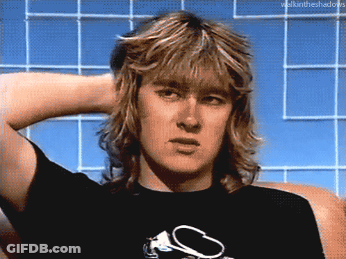 A Man With Long Blonde Hair Is Wearing A Black T-Shirt With A S On It GIF