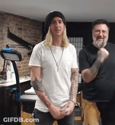 A Man With Long Blonde Hair Stands Next To Another Man With A Beard GIF