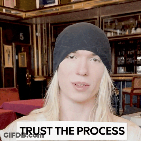 A Man With Long Blonde Hair Wearing A Black Hat With A Sign That Says Trust The Process GIF