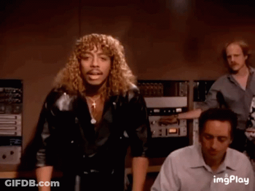A Man With Long Curly Hair And A Mustache Is Dancing In A Room . GIF