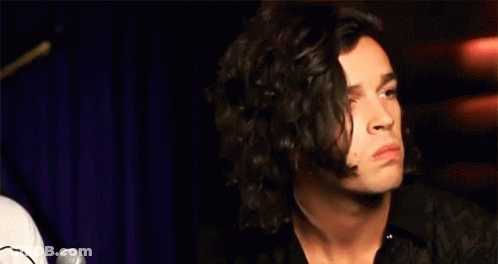 A Man With Long Curly Hair Is Sitting In Front Of A Microphone Making A Funny Face . GIF