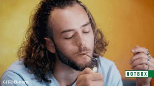A Man With Long Curly Hair Is Smoking A Cigarette In Front Of A Hotbox Sign GIF