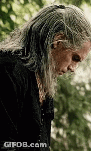 A Man With Long Gray Hair And A Black Shirt Is Standing In A Forest . GIF