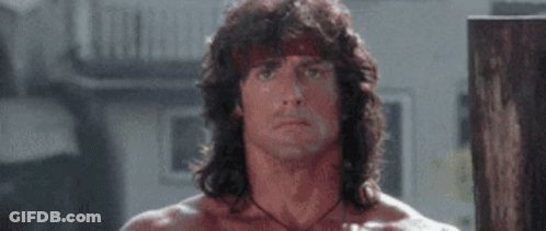A Man With Long Hair And A Bandana On His Head Is Standing In Front Of A Wooden Wall . GIF
