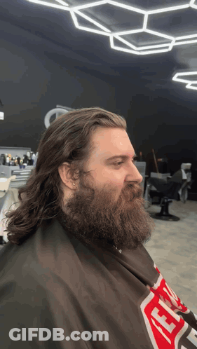 A Man With Long Hair And A Beard Is Getting His Hair Cut By A Barber Wearing A Supreme Cape GIF