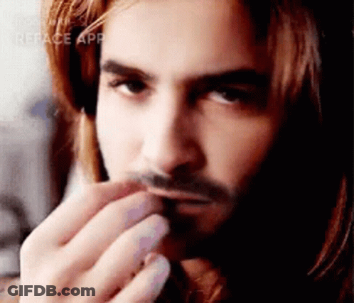 A Man With Long Hair And A Beard Is Holding His Hand To His Mouth . GIF