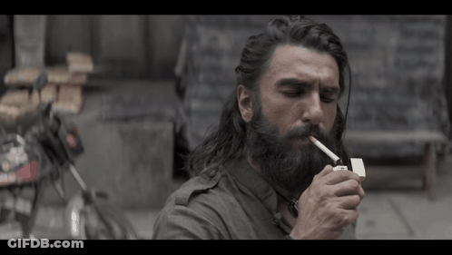 A Man With Long Hair And A Beard Is Lighting A Cigarette With A Lighter GIF