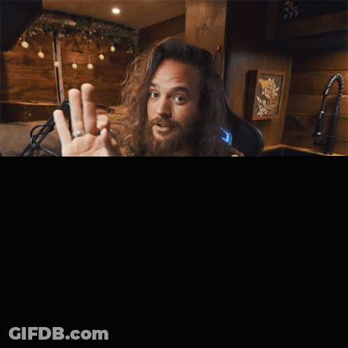 A Man With Long Hair And A Beard Is Sitting At A Desk With The Words So Listen Up Behind Him GIF