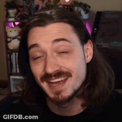 A Man With Long Hair And A Beard Is Smiling In Front Of A Bookshelf . GIF