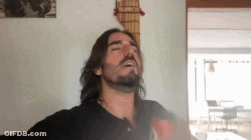 A Man With Long Hair And A Beard Is Smoking A Cigarette In Front Of A Guitar Hanging On A Wall . GIF