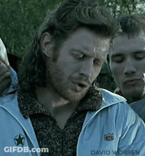 A Man With Long Hair And A Beard Is Smoking A Cigarette While Standing Next To Another Man . GIF