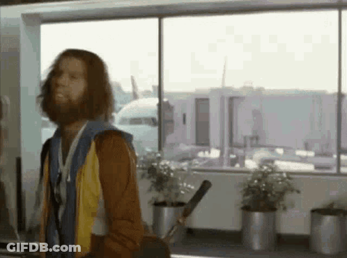 A Man With Long Hair And A Beard Is Standing In Front Of A Window Holding A Golf Club . GIF