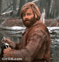 A Man With Long Hair And A Beard Is Standing In The Woods Holding A Gun GIF