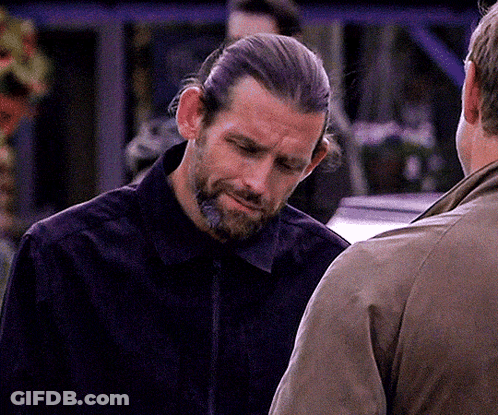 A Man With Long Hair And A Beard Is Talking To Another Man In A Brown Jacket GIF