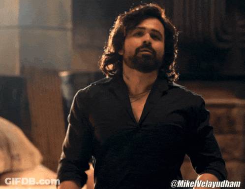 A Man With Long Hair And A Beard Is Wearing A Black Shirt With @Mikelvelayudham On The Bottom GIF