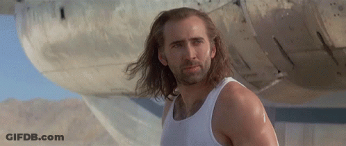 A Man With Long Hair And A Beard Wearing A White Tank Top Is Standing In Front Of A Plane . GIF