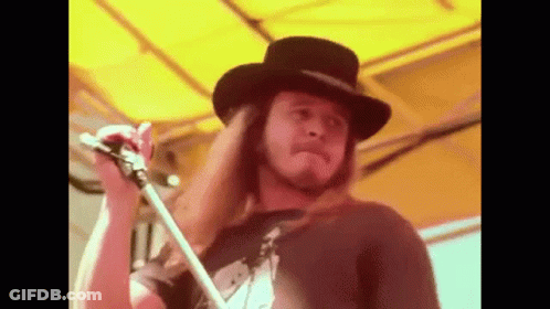A Man With Long Hair And A Hat Is Holding A Microphone . GIF