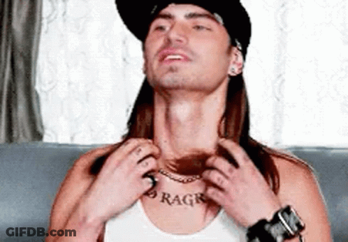 A Man With Long Hair And A Hat Is Sitting On A Couch Adjusting His Necklace . GIF
