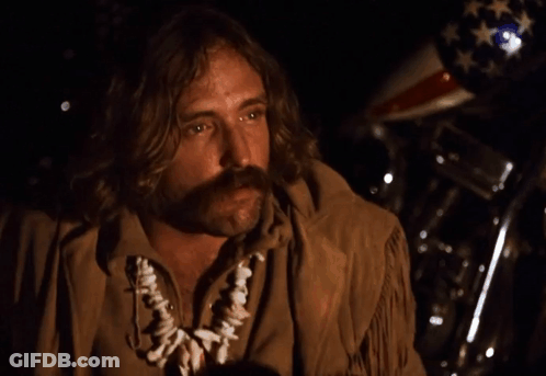 A Man With Long Hair And A Mustache Is Standing In Front Of A Motorcycle With An American Flag In The Background GIF