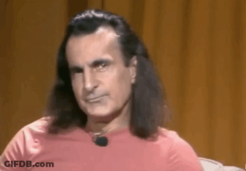 A Man With Long Hair And A Pink Shirt Is Sitting In Front Of A Brown Curtain . GIF