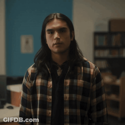 A Man With Long Hair And A Plaid Shirt Looks At The Camera GIF