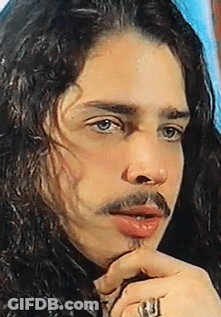 A Man With Long Hair And A Ring On His Finger Looks At The Camera GIF