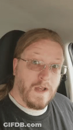 A Man With Long Hair And Glasses Is Sitting In A Car And Making A Funny Face . GIF