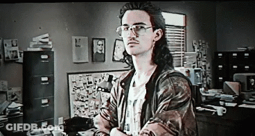 A Man With Long Hair And Glasses Is Standing In Front Of A Desk In An Office . GIF