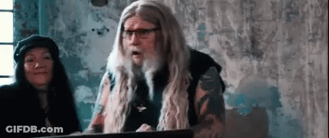 A Man With Long Hair And Glasses Is Standing In Front Of A Podium . GIF