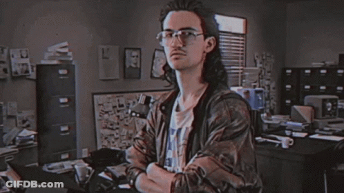 A Man With Long Hair And Glasses Is Standing In Front Of A Sign That Says Hackerman GIF