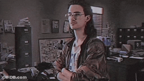 A Man With Long Hair And Glasses Stands In Front Of A Sign That Says Hackerman GIF