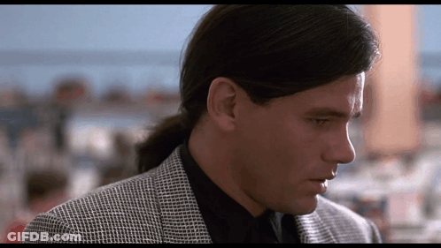 A Man With Long Hair In A Ponytail Is Wearing A Black Shirt And A Grey Jacket GIF
