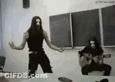 A Man With Long Hair Is Dancing In Front Of A Blackboard In A Classroom While Another Man Plays A Guitar . GIF