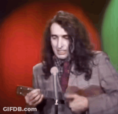 A Man With Long Hair Is Holding A Guitar And Speaking Into A Microphone . GIF
