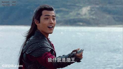A Man With Long Hair Is Holding A Rock In Front Of A Body Of Water And Has Chinese Writing On His Arm GIF