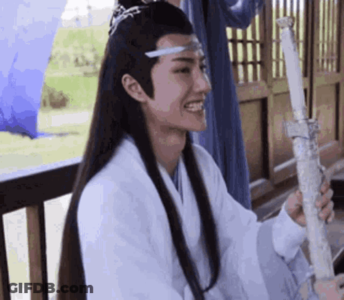A Man With Long Hair Is Holding A Sword And Smiling While Sitting On A Balcony . GIF