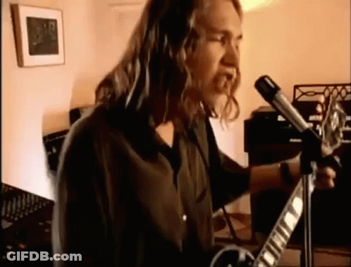 A Man With Long Hair Is Playing A Guitar In Front Of A Microphone In A Room . GIF