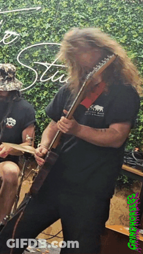 A Man With Long Hair Is Playing A Guitar In Front Of A Sign That Says Stu GIF