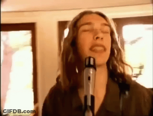 A Man With Long Hair Is Singing Into A Microphone In A Room . GIF
