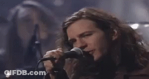 A Man With Long Hair Is Singing Into A Microphone While Another Man Looks On . GIF