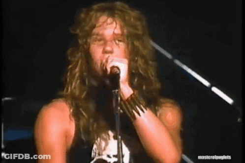 A Man With Long Hair Is Singing Into A Microphone While Standing On A Stage . GIF