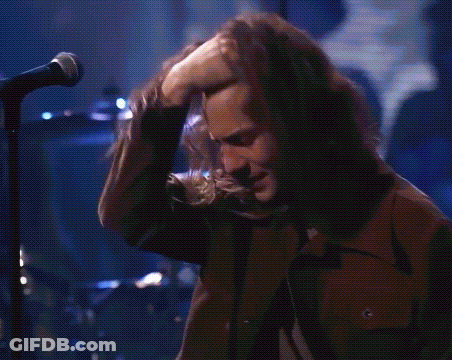 A Man With Long Hair Is Singing Into A Microphone While Touching His Nose . GIF