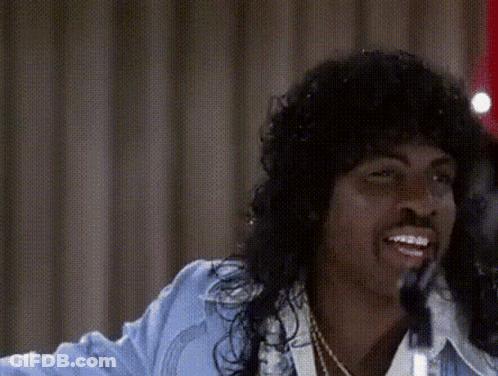A Man With Long Hair Is Singing Into A Microphone While Wearing A Blue Jacket . GIF