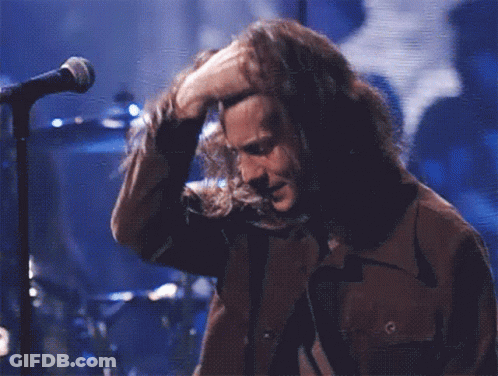 A Man With Long Hair Is Singing Into A Microphone While Wearing A Brown Jacket GIF