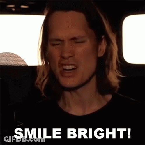 A Man With Long Hair Is Singing Into A Microphone With The Words Smile Bright Behind Him GIF