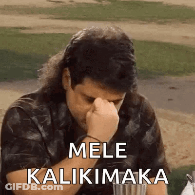 A Man With Long Hair Is Sitting At A Table With His Hand On His Forehead And The Words Mele Kalikimaka On The Screen . GIF
